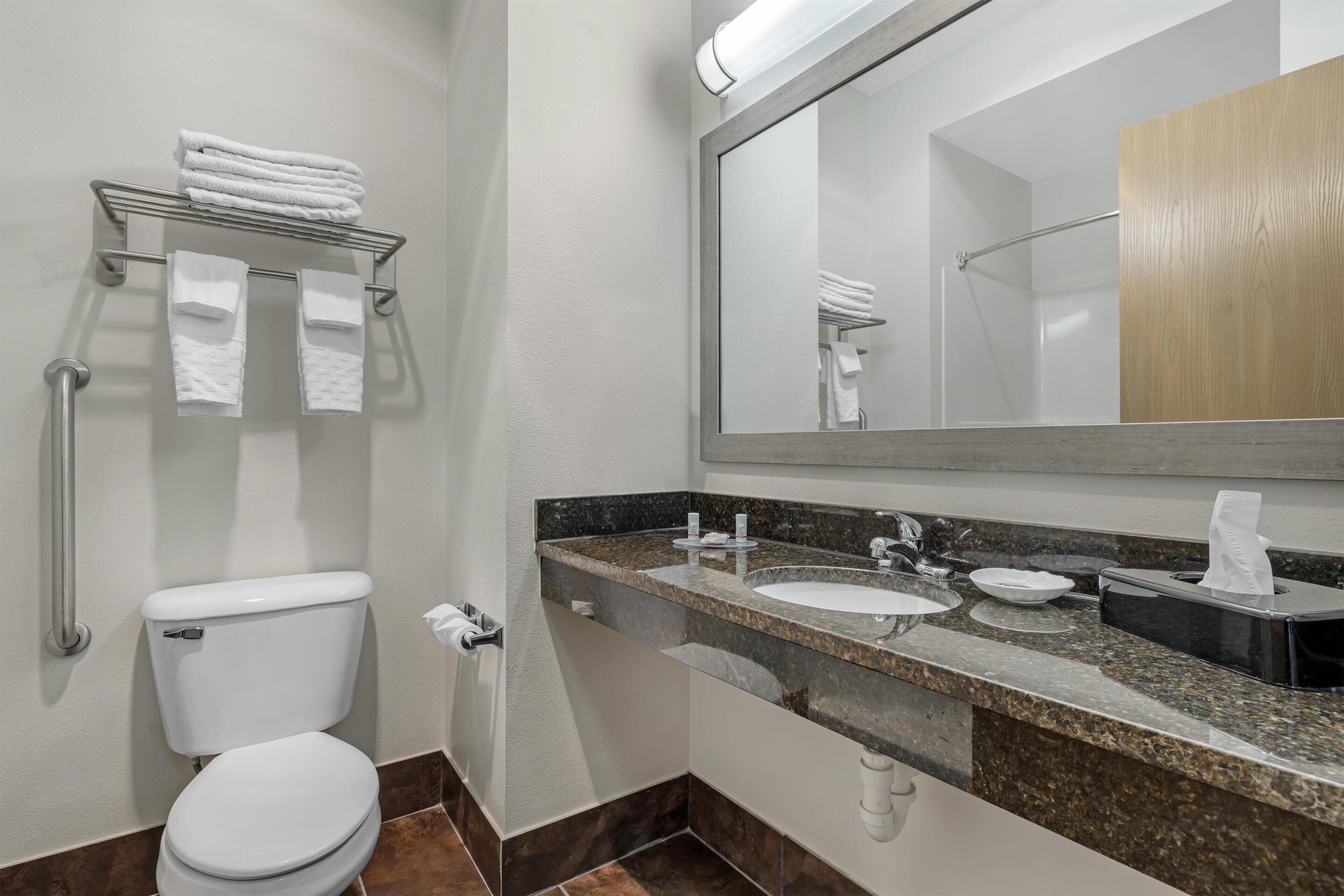 A bathroom with a toilet, sink, mirror, towel rack, and a towel dispenser.