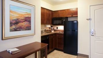 A compact kitchen with wooden cabinets, a black refrigerator, a microwave, and a sink. There's a framed painting on the wall and a small table with a book.