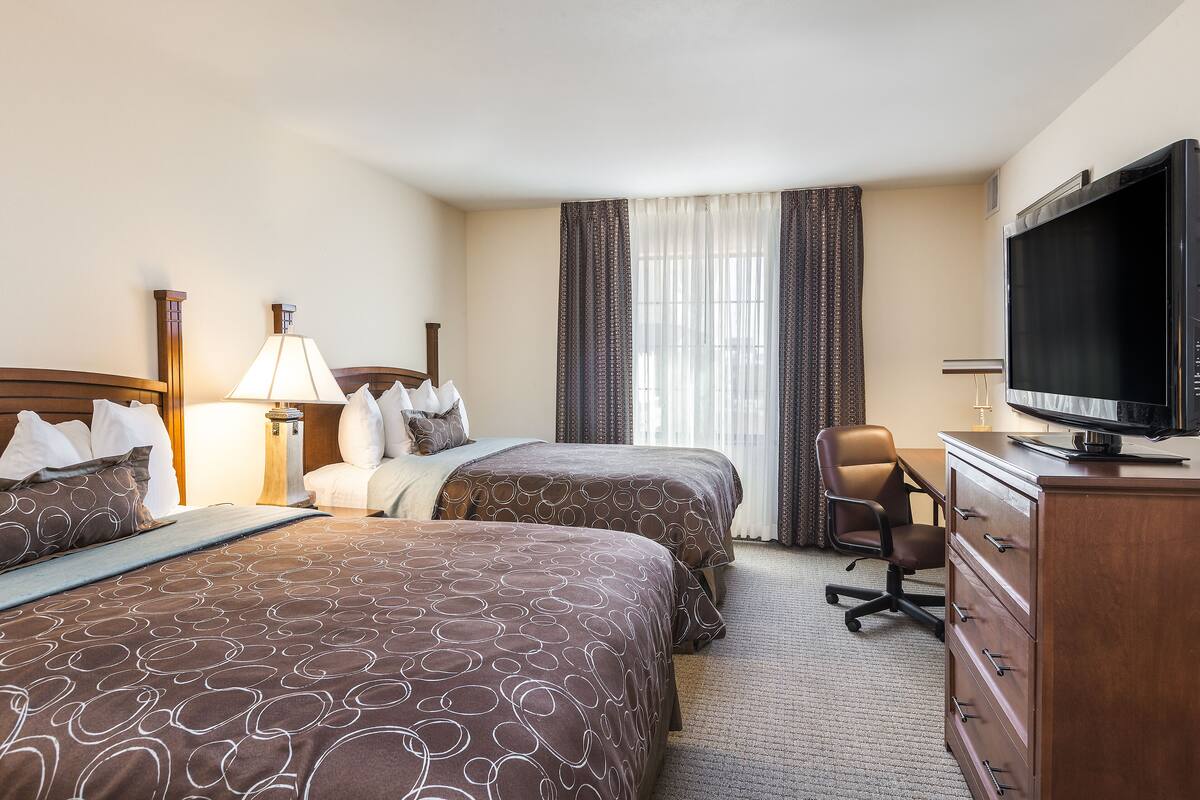Quarto de hotel com duas camas, uma escrivaninha, uma cadeira, uma televisão e uma luminária.
