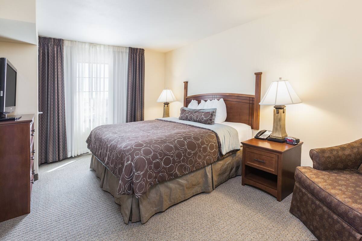 Quarto de hotel com uma cama grande, duas luminárias de cabeceira, uma poltrona, uma televisão e uma escrivaninha com luminária.