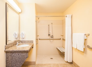 Baño accesible para personas con movilidad reducida, con lavamanos de encimera de granito, un espejo, ducha con barras de apoyo y una toalla blanca colgada en un perchero.
