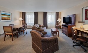 A hotel room with a living area, a dining table with chairs, a sofa, a TV, and a desk with a lamp.