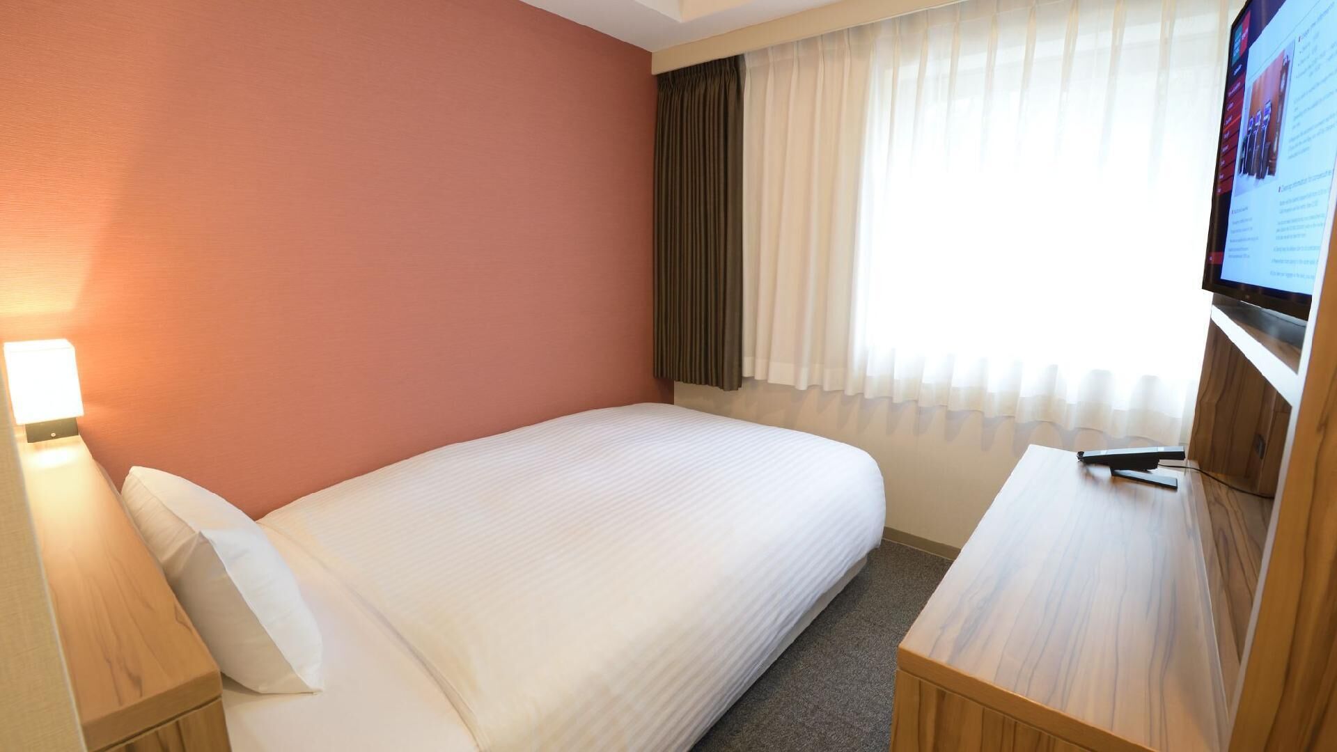 A hotel room with a single bed, a desk, a TV, and a window with curtains.