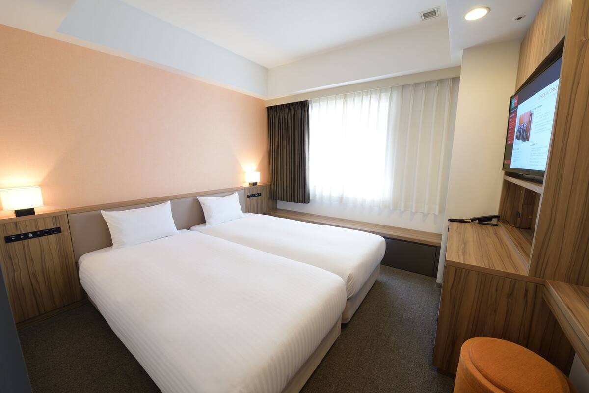 A hotel room with two beds, a TV, a desk, and a window with curtains.