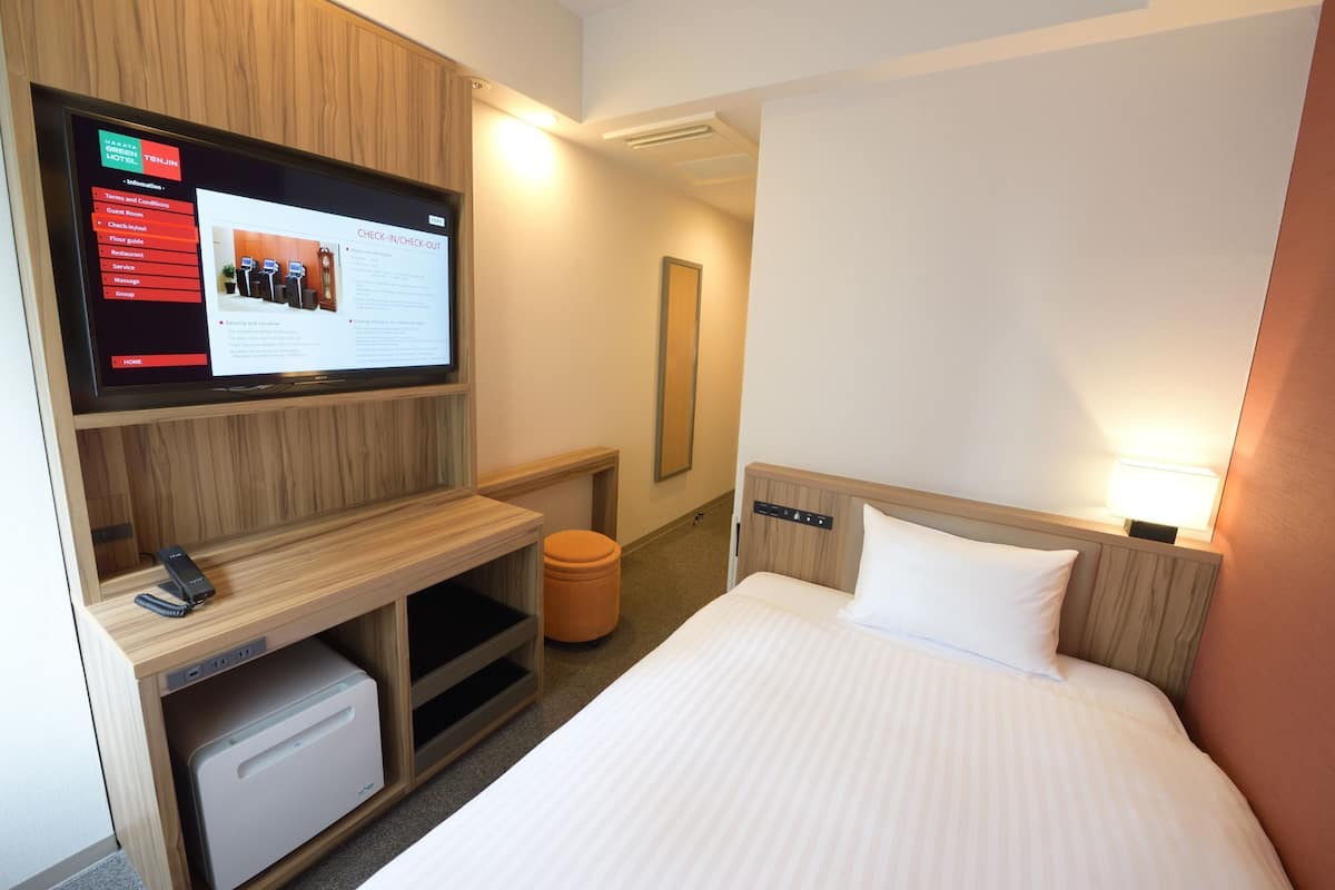 A hotel room with a bed, a desk with a computer, a TV mounted on the wall, and a small trash bin.