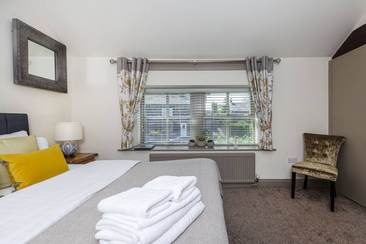 A hotel room with a bed, a chair, a window with curtains, a radiator, and a mirror on the wall.