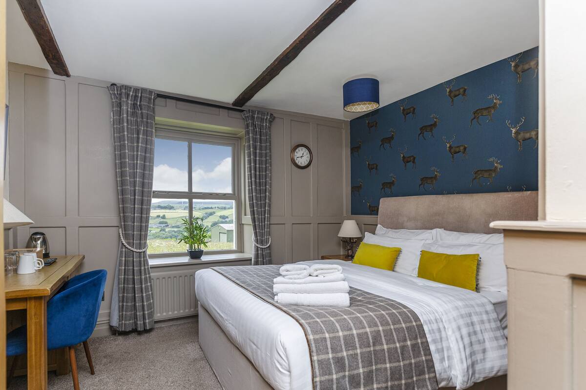 A hotel room with a bed, a desk, a chair, a window with curtains, a clock, and a wall with a deer pattern.