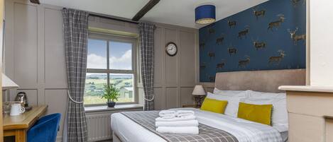 A hotel room with a bed, a desk, a chair, a window with curtains, a clock, and a wall with a deer pattern.