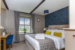 Una camera d'albergo con un letto, una scrivania, una sedia, una finestra con tende, una sveglia e una parete decorata con un motivo cervi.
