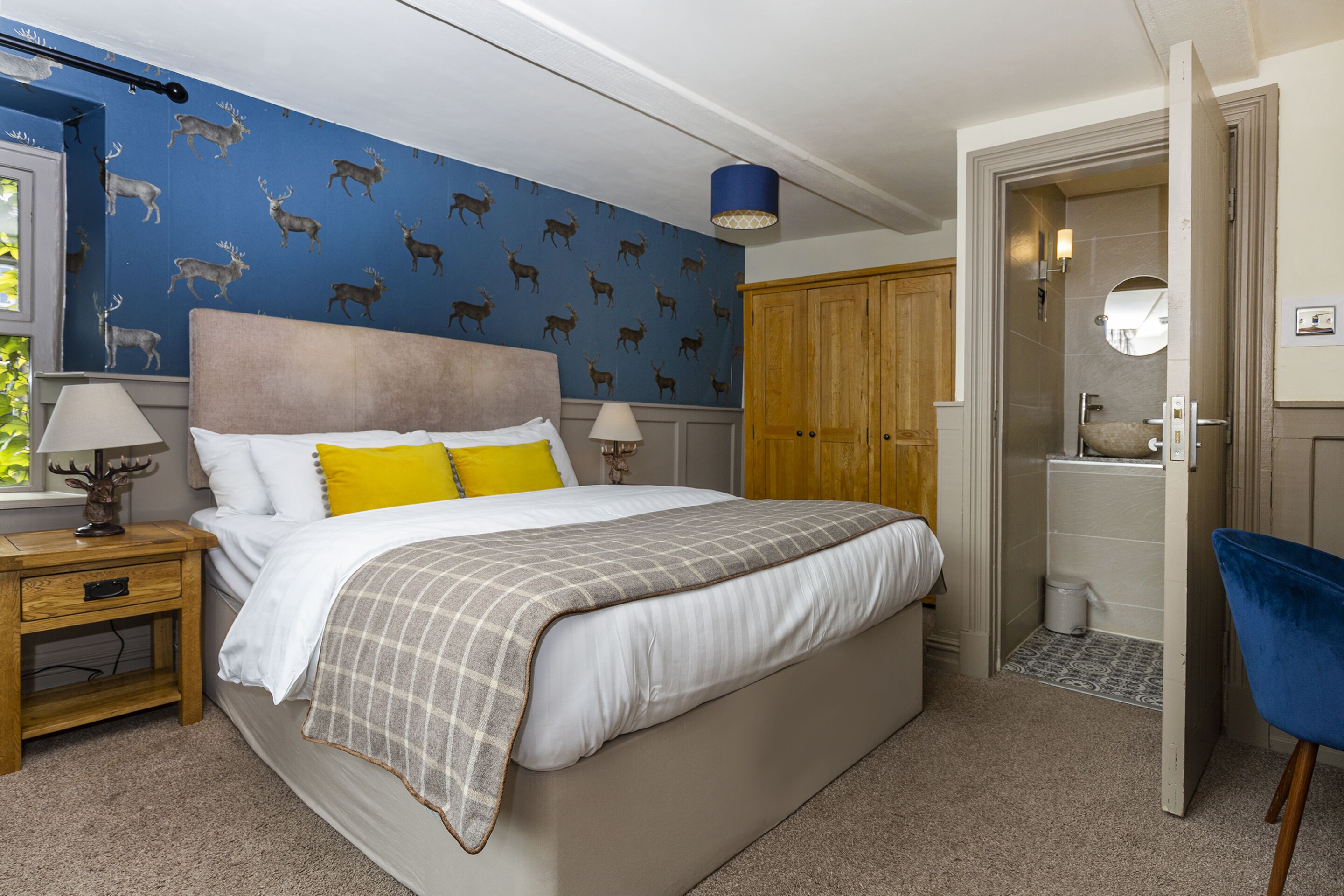 A bedroom with a bed, nightstands, a blue wall with deer wallpaper, a wooden wardrobe, and a bathroom visible through an open door.