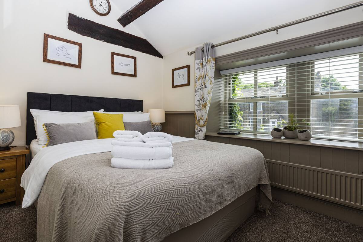A neatly made bed with a headboard, a bedside table with a lamp, and a window with blinds and plants.