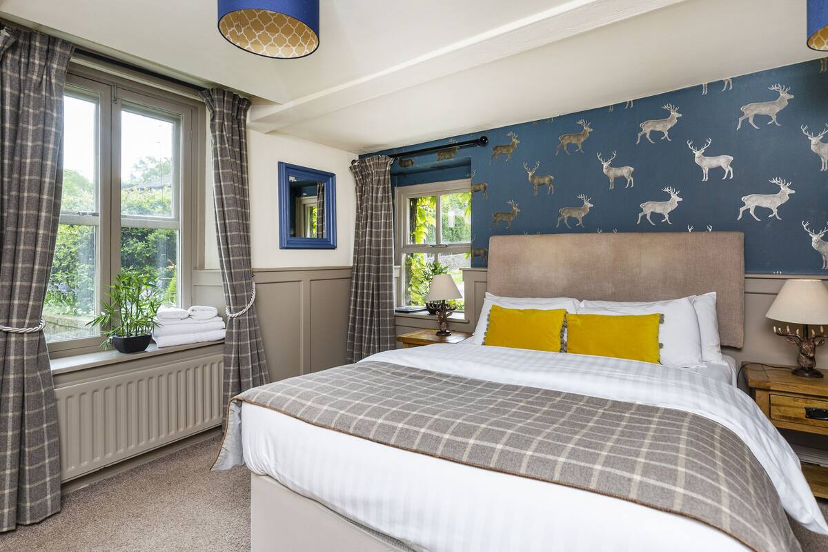 A bedroom with a large bed, a window with curtains, a radiator, a nightstand with a lamp, and a wall with deer wallpaper.