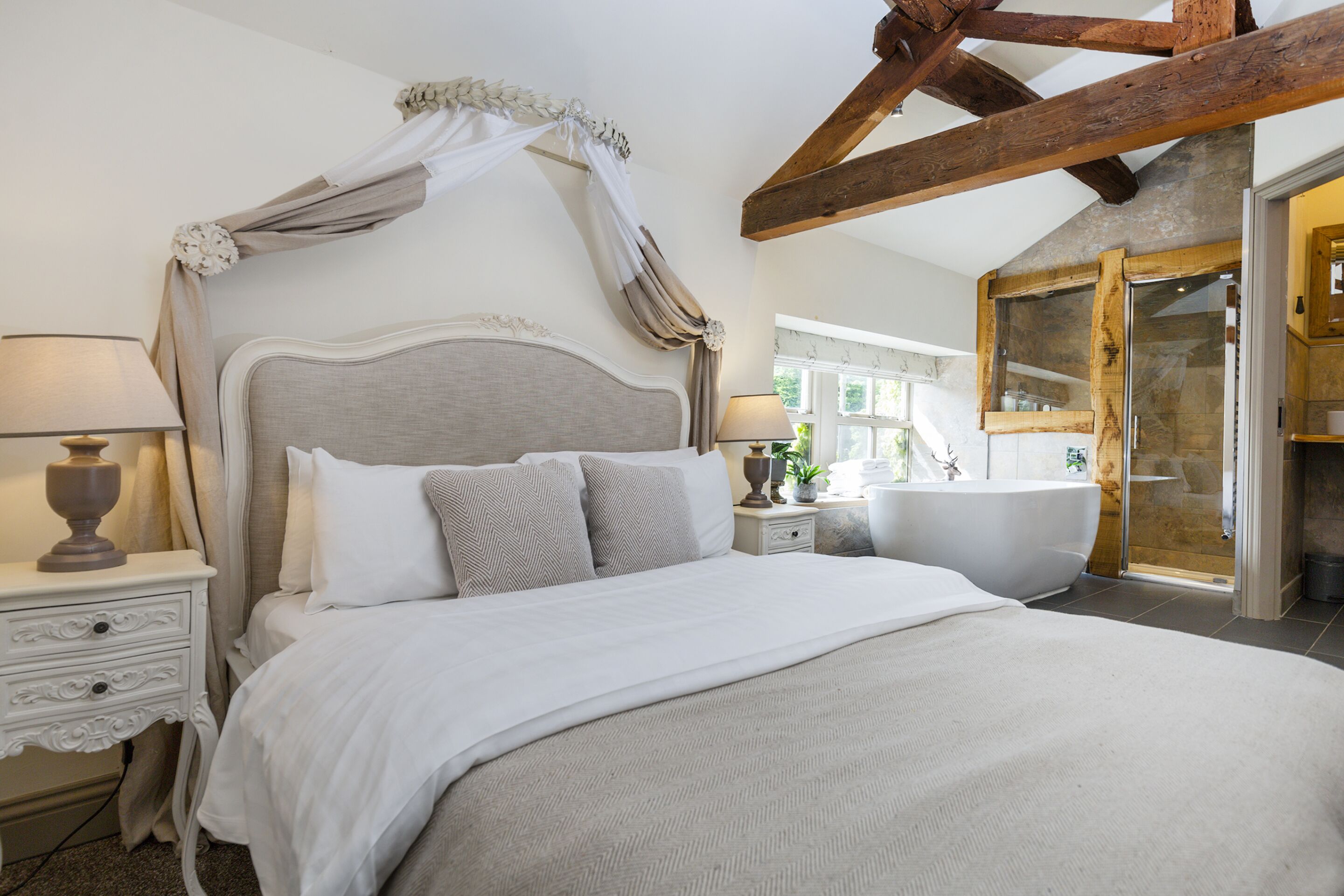 A bedroom with a large bed, a nightstand, a lamp, a window, and a bathroom with a bathtub.