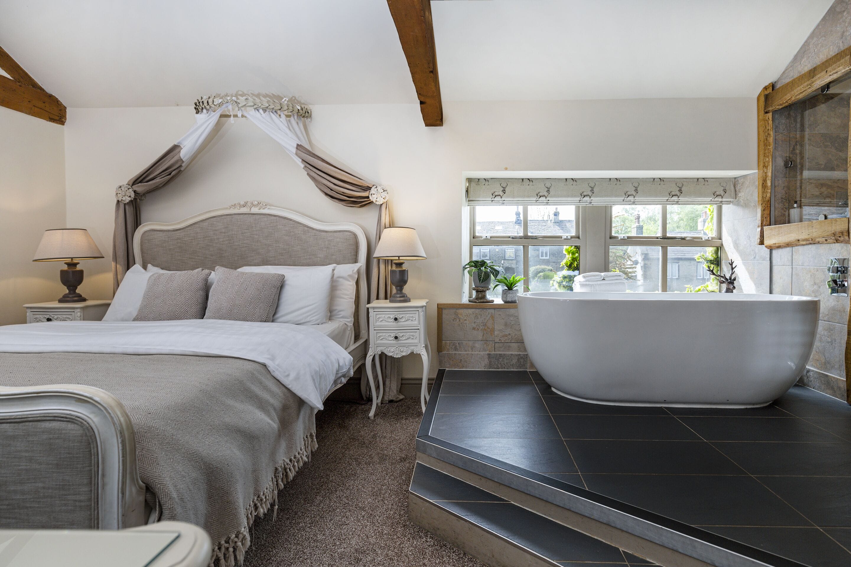 A bedroom with a large bed, a canopy, a nightstand with a lamp, a window with a view of buildings, and a bathtub.