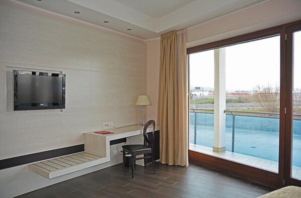 Una habitación de hotel moderna con un televisor de pantalla plana montado en la pared, un escritorio con una silla y un ventanal con vistas a una piscina y edificios.
