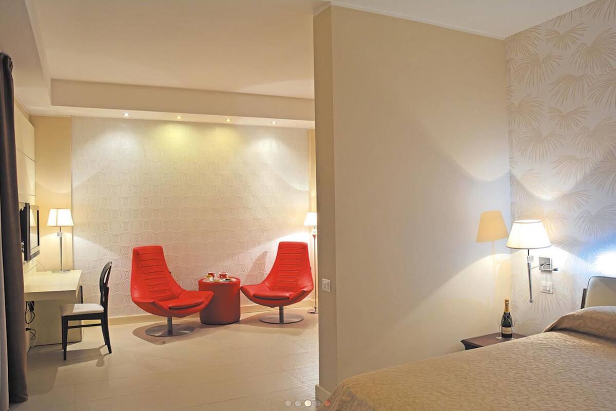 A hotel room with a bed, two red chairs, a small table, a desk, and a nightstand with a lamp and a bottle.
