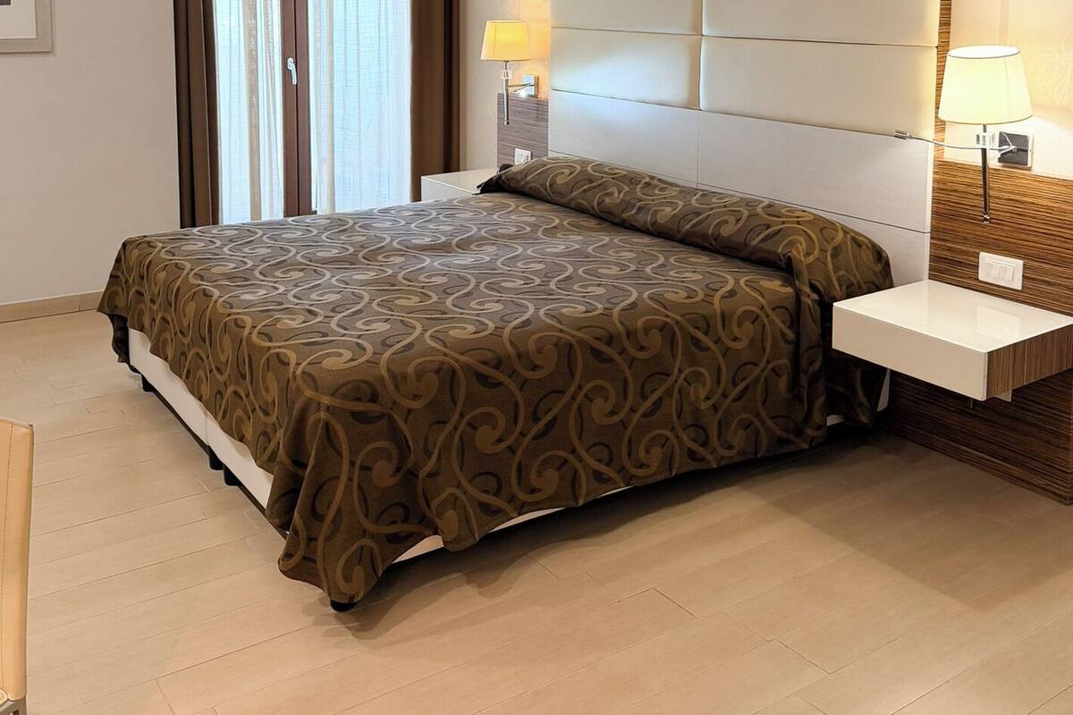 A hotel room with a large bed, two bedside tables, a headboard, a door with curtains, and a light-colored floor.
