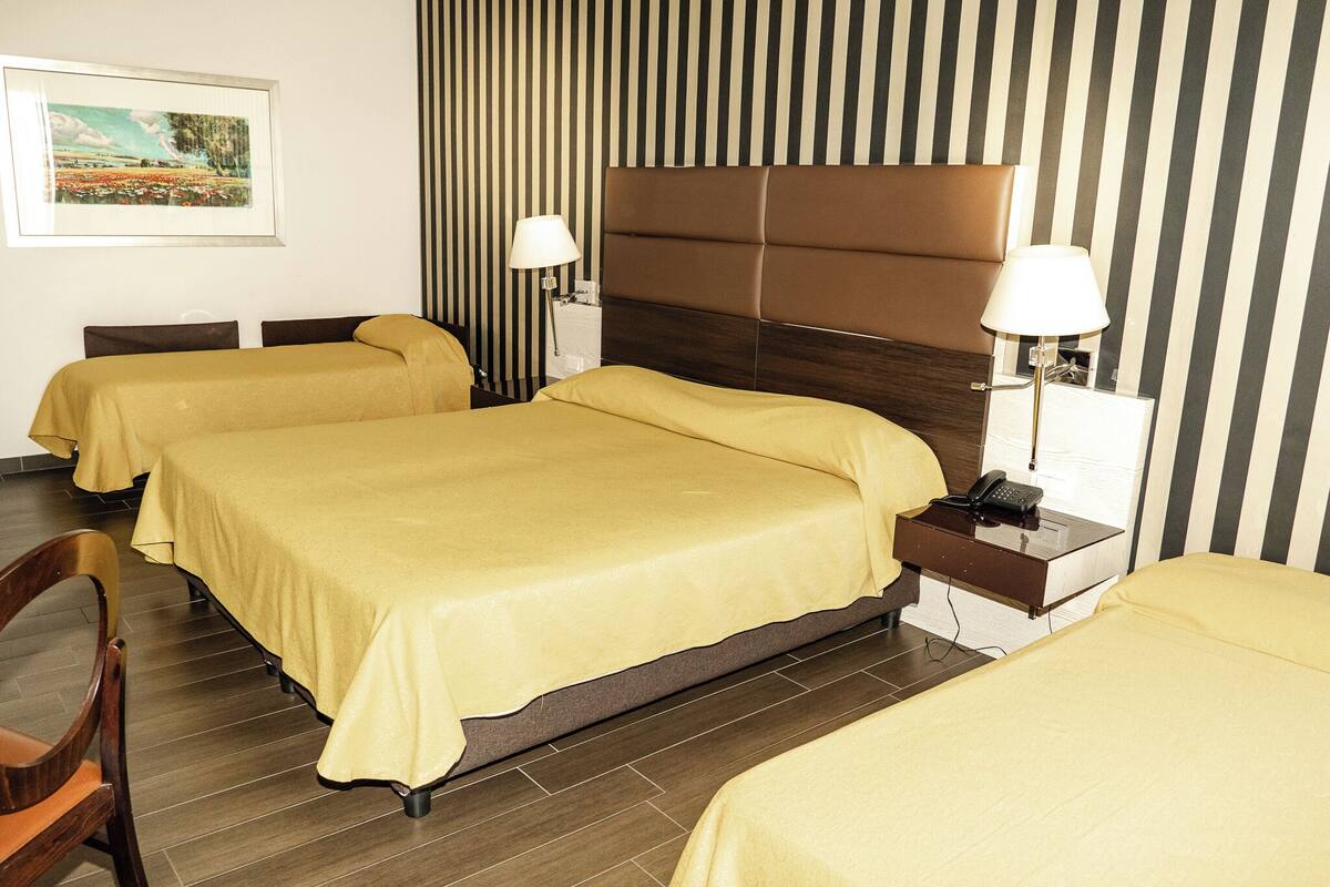 A hotel room with three beds, each with a yellow bedspread, a wooden headboard, and a nightstand with a lamp and phone.