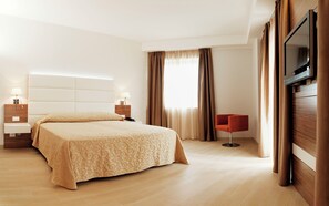Una habitación de hotel con una cama grande, una mesita de noche con una lámpara, un televisor montado en la pared y una silla.