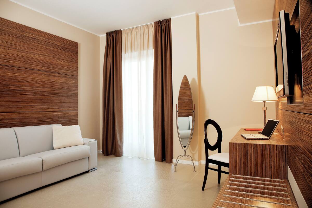 A hotel room with a sofa, a desk with a laptop, a mirror, and a TV on the wall.