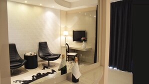 Una habitación de hotel moderna con un espejo grande, dos sillones de cuero negro, una mesita con un jarrón y flores, una botella de champán y un televisor de pantalla plana sobre un estante blanco.
