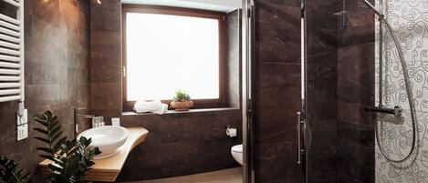 A modern bathroom with a glass-enclosed shower, a large window, a sink with a marble countertop, and a potted plant.
