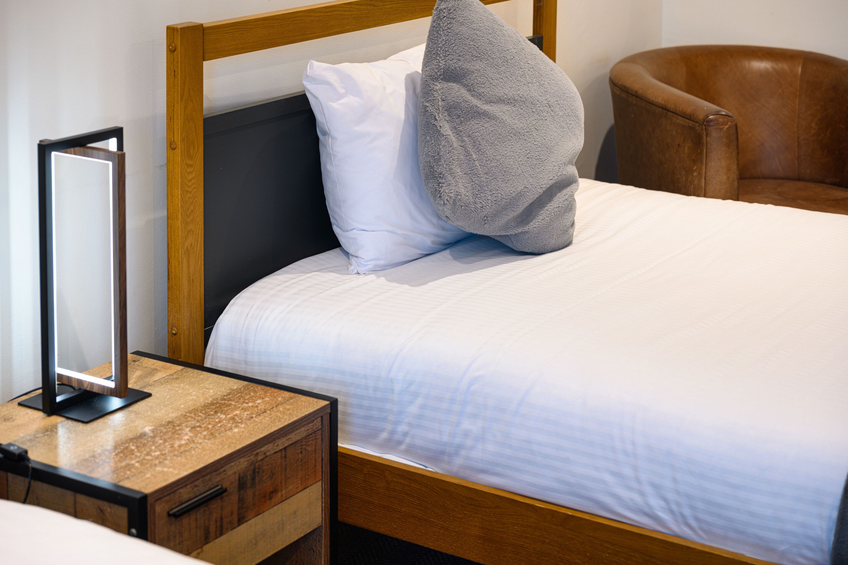 Una cama bien hecha con almohadas blancas y grises, un cabecero de madera y un sillón de cuero marrón.