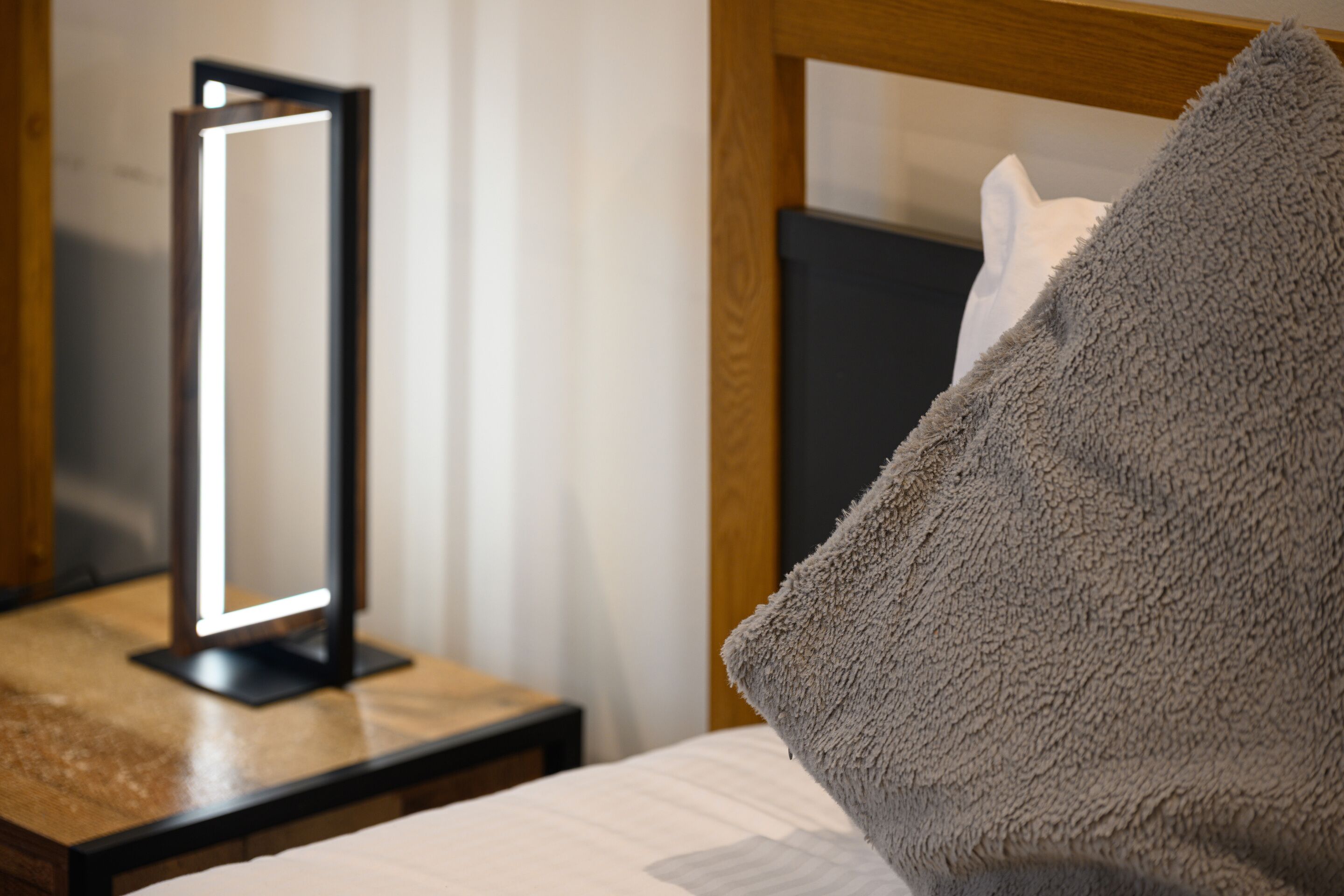 A neatly made bed with a gray blanket, a bedside table with a modern lamp, and a wooden headboard.