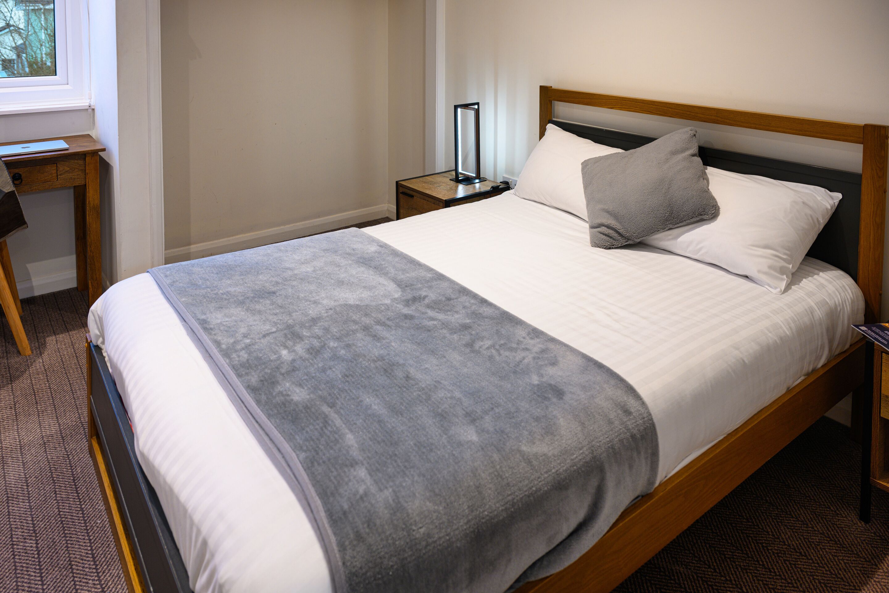 Una cama bien tendida con una manta gris, un cabecero de madera y una mesita de noche con una lámpara.