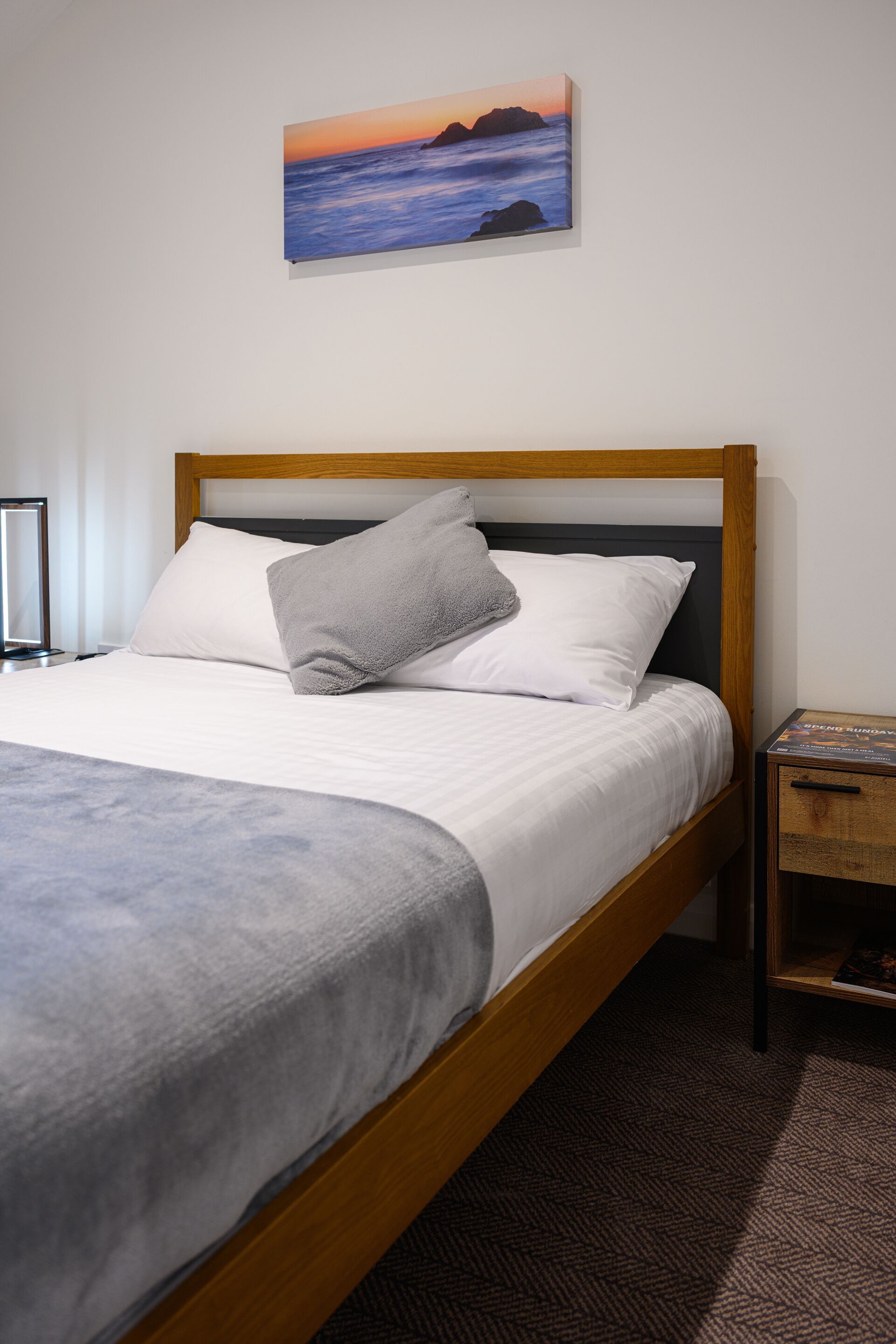 A neatly made bed with a wooden headboard, a gray throw blanket, and a pillow. A framed painting of a sunset over the ocean hangs above the bed.