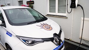 Een witte auto geparkeerd voor een gebouw met een bord waarop staat 'Delicious Specials' en een laadstation voor elektrische voertuigen dat aan de muur is bevestigd.