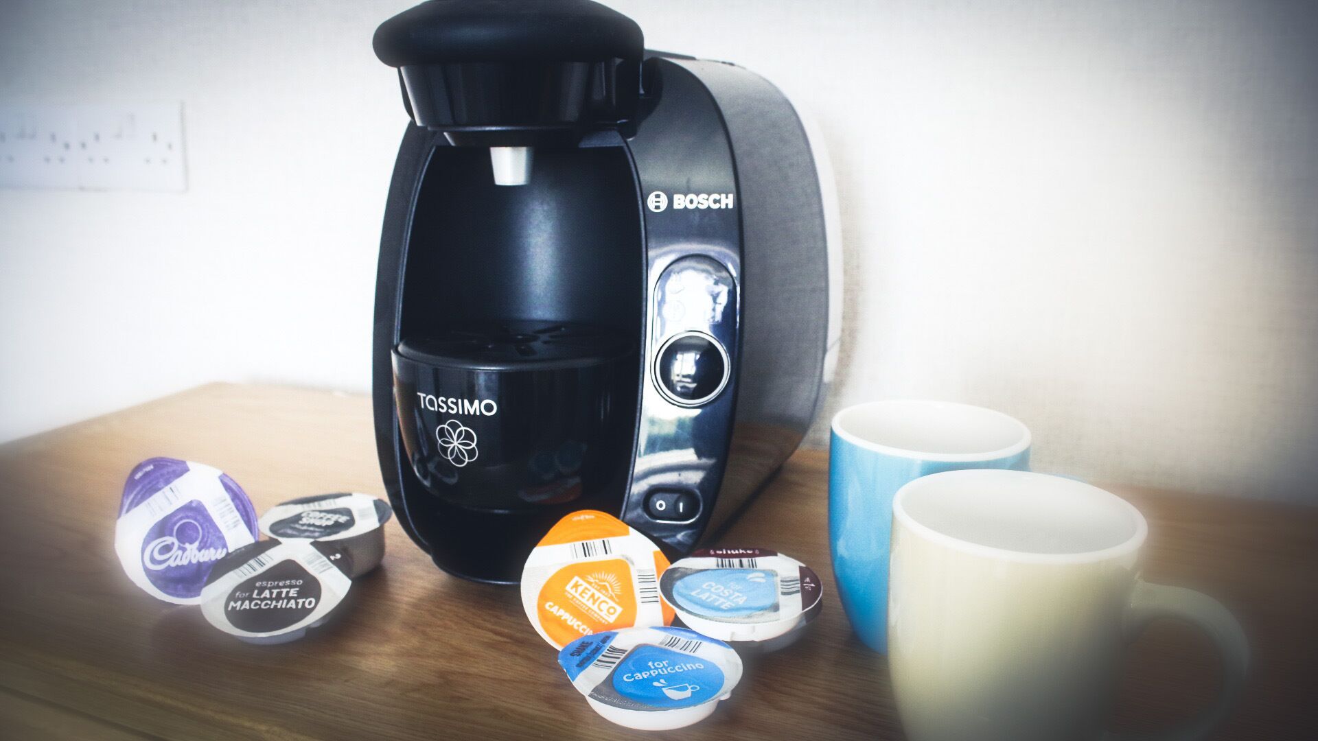 A coffee maker, coffee pods, and two mugs on a table.