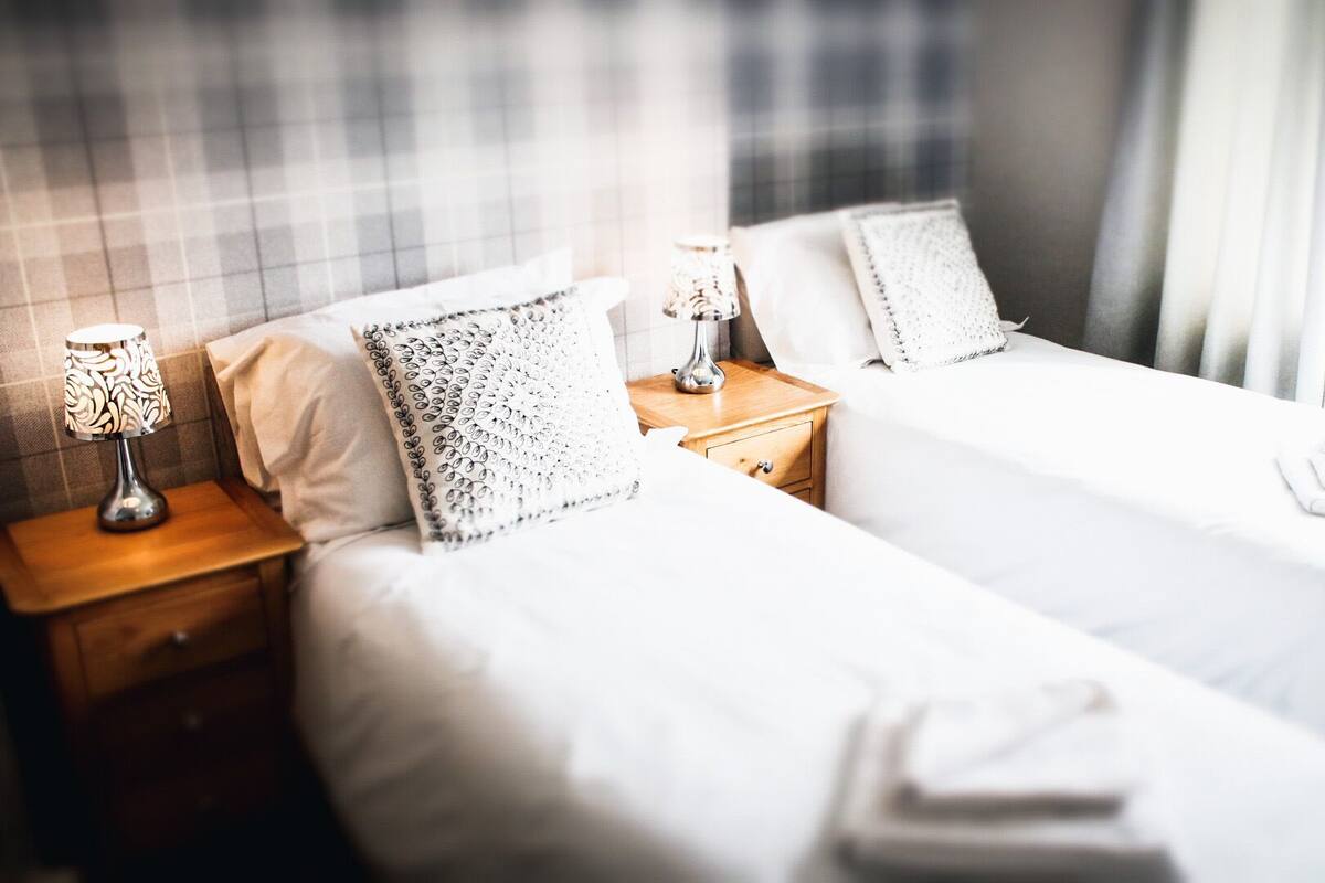 A hotel room with two beds, each with white linens and decorative pillows. There are bedside tables with lamps and a window with curtains.