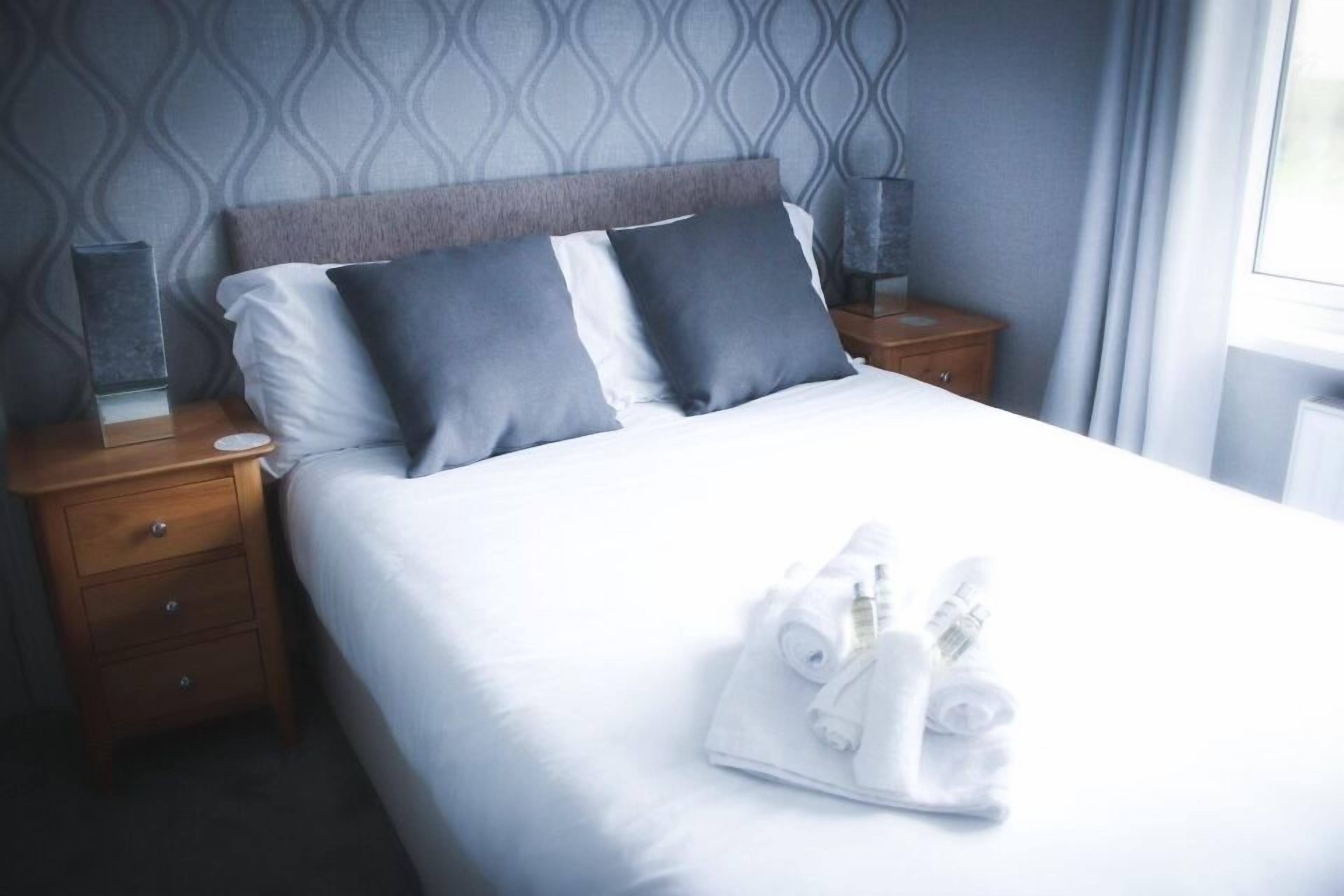 Una cama bien hecha con sábanas blancas y dos almohadas oscuras, una mesita de noche de madera con una lámpara, y un juego de toallas sobre la cama.