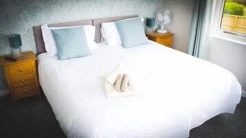 Una cama bien tendida con sábanas blancas y dos almohadas azul claro, una mesita de noche de madera con una lámpara, y una ventana con vistas a follaje.