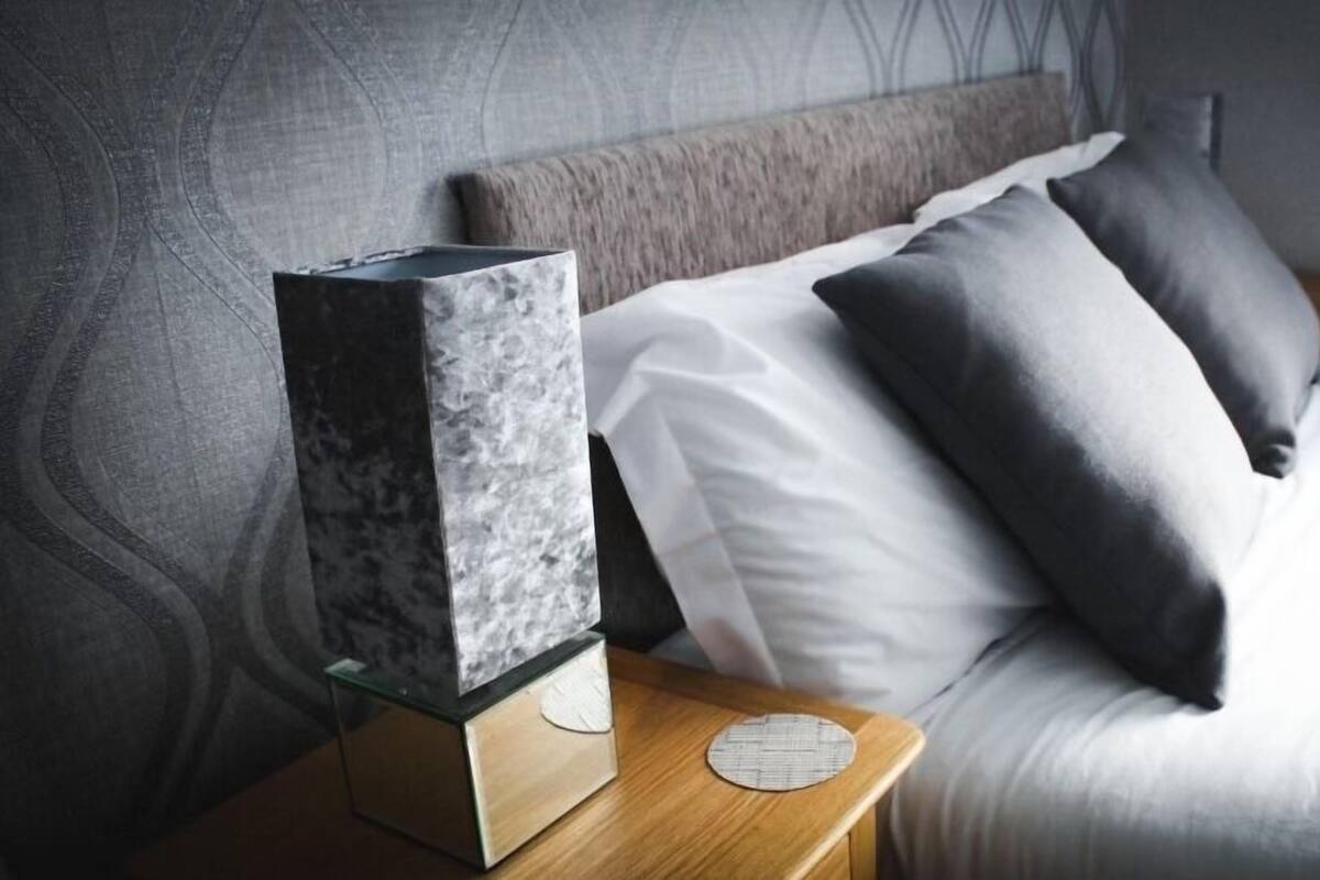 A neatly made bed with white and grey bedding, a marble-like object on a wooden nightstand, and a patterned round coaster.
