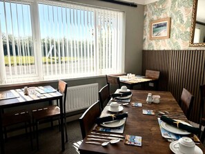 A dining area with tables set for two, a view of the outdoors through large windows, and a tropical wallpaper design. - Sandyhill Guest House (Saundersfoot)