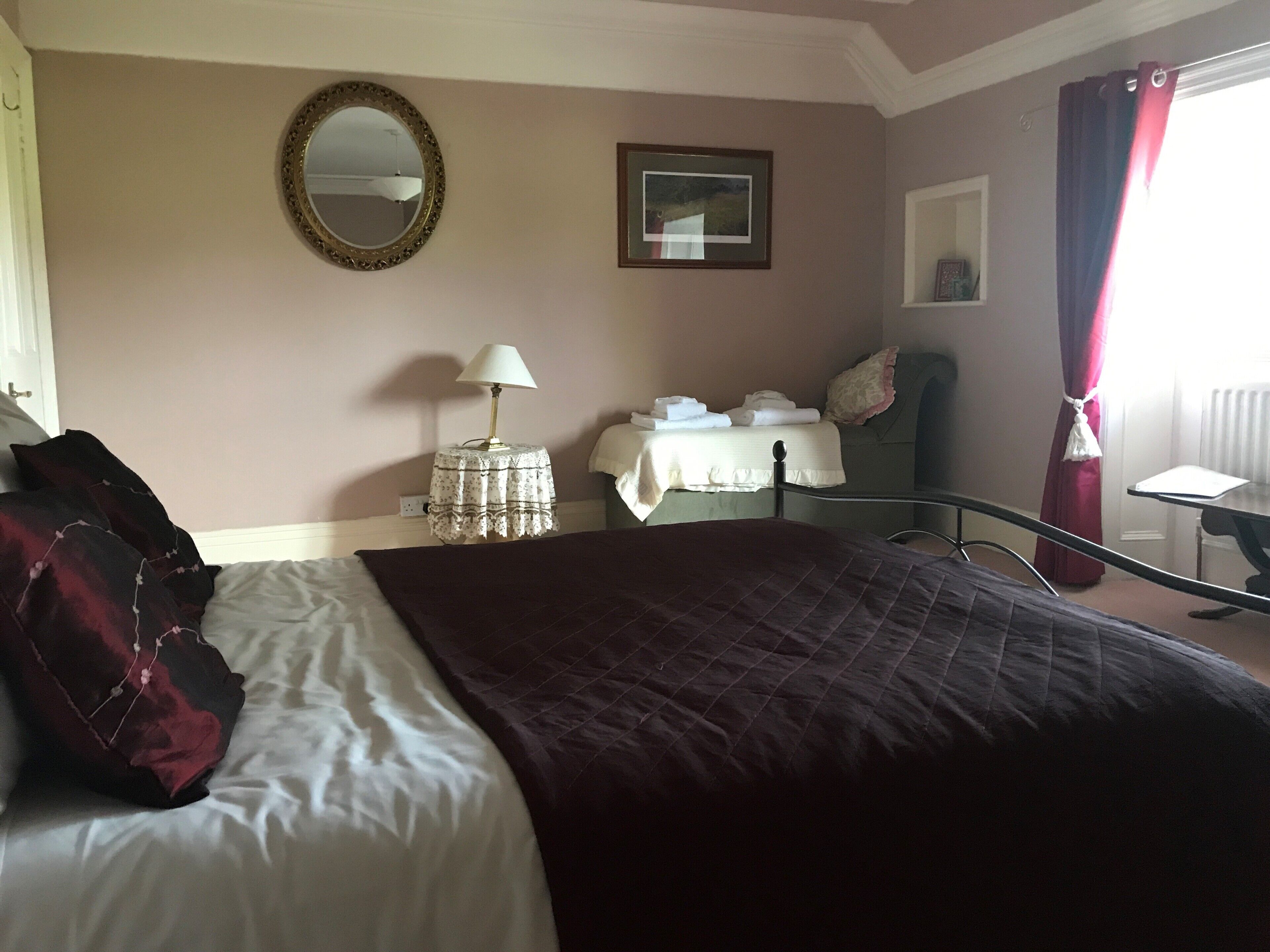 A hotel room with a large bed, a bedside table with a lamp, a small table with towels, a chair, and a window with curtains.