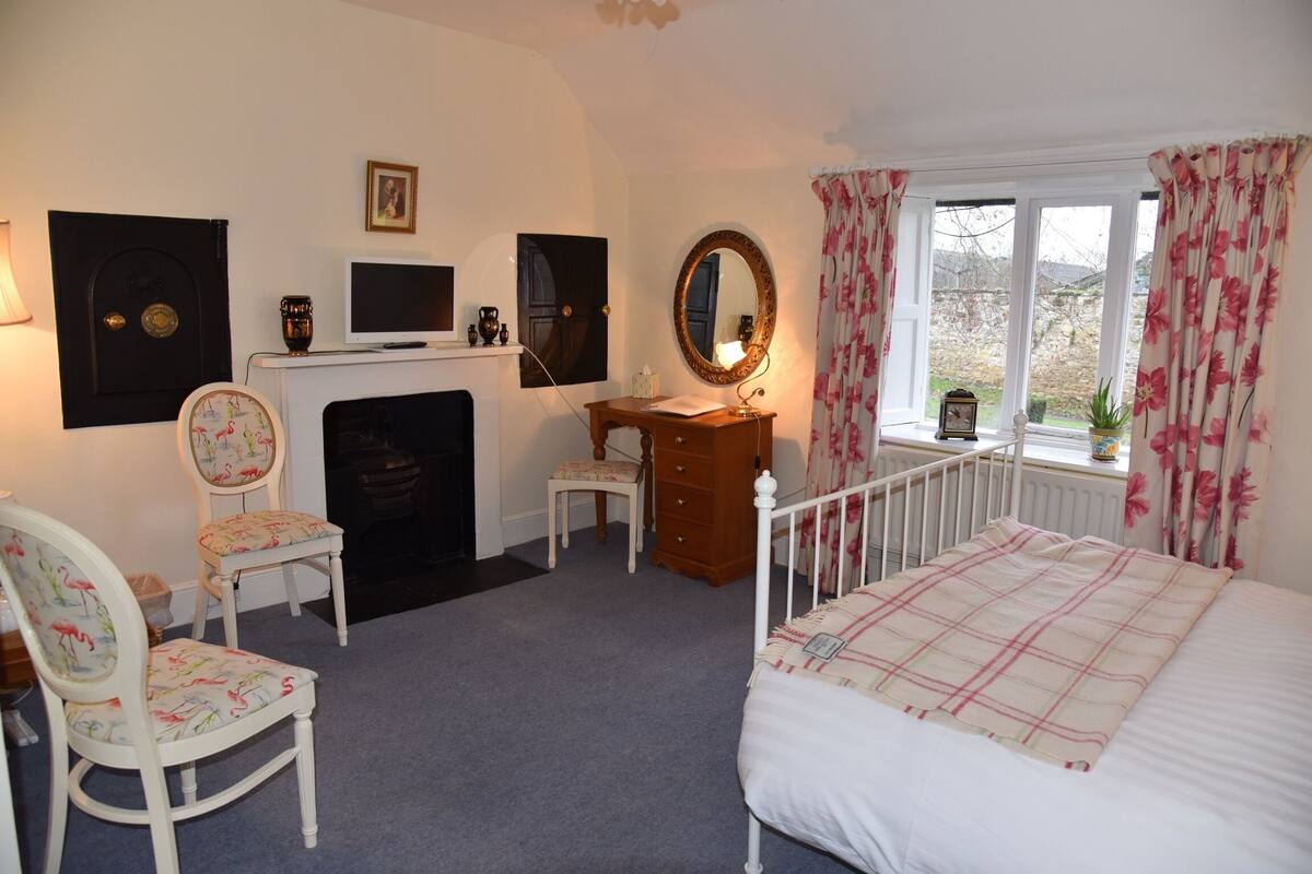 A bedroom with a bed, a desk, a chair, a dresser, a fireplace, and a window with curtains.