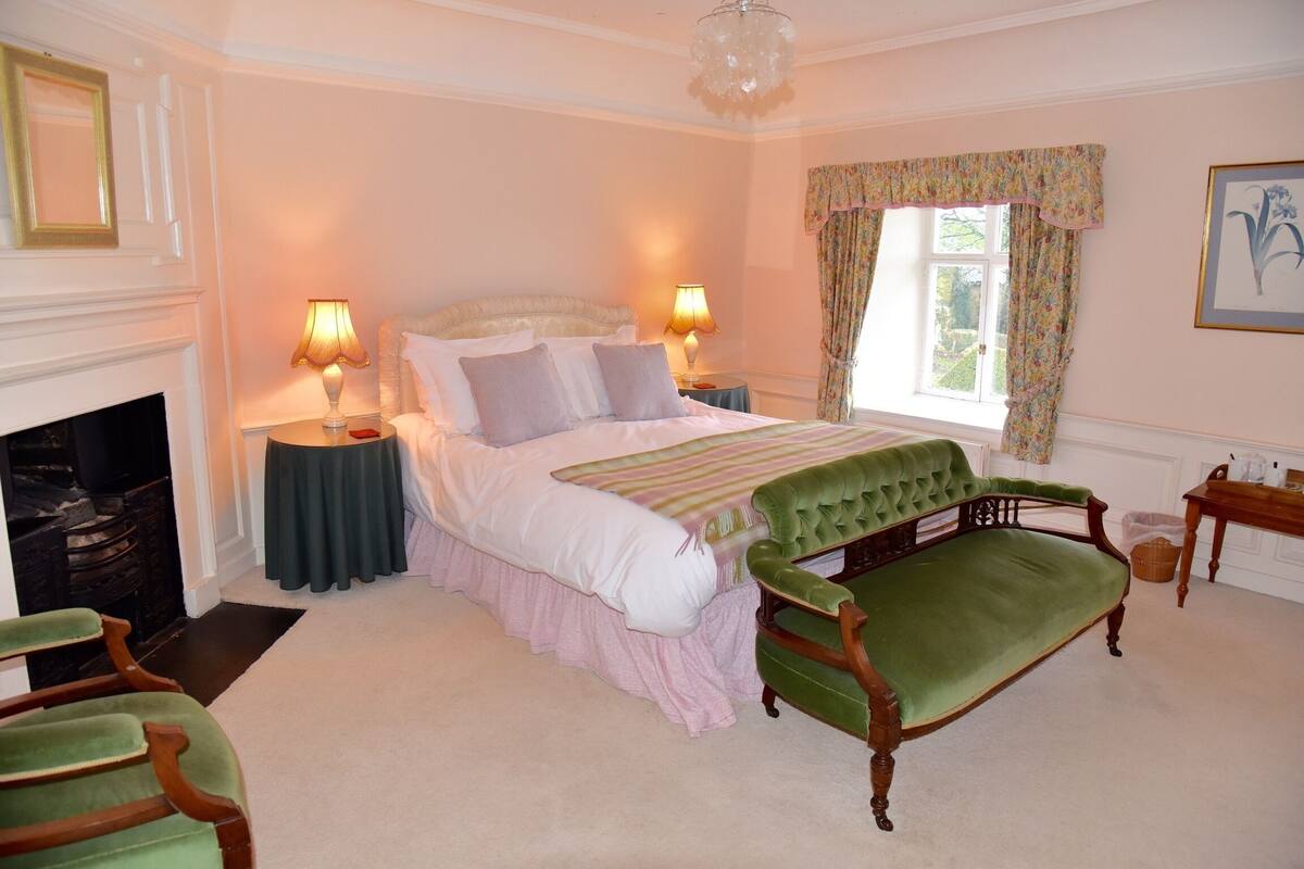A bedroom with a bed, two bedside lamps, a green upholstered bench, a fireplace, and a window with floral curtains.