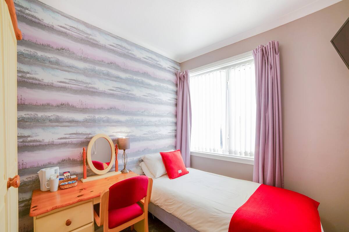 A bedroom with a bed, a desk with a chair, a mirror, and a window with curtains.