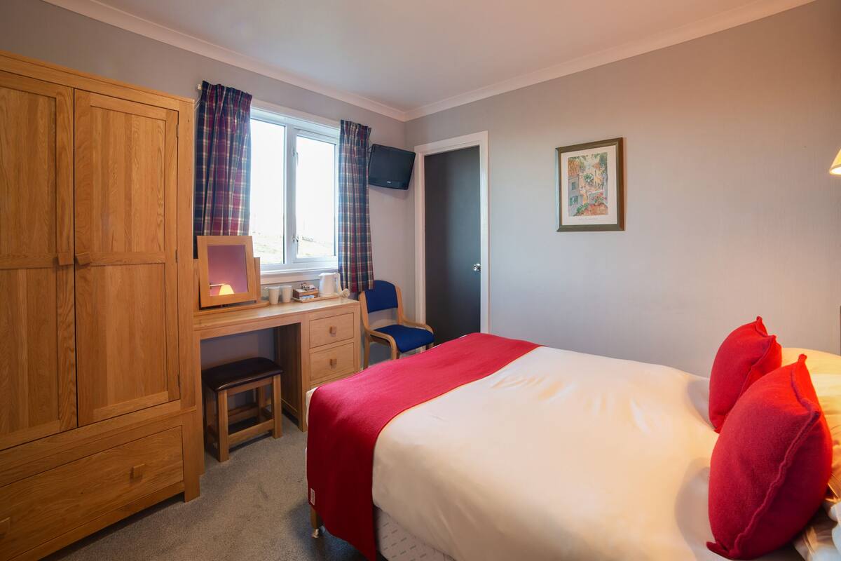 A bedroom with a bed, a desk with a chair, a wardrobe, a window with curtains, and a door.