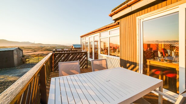 Eine Holzterrasse mit einem weißen Tisch und Stühlen, mit Blick auf eine malerische Landschaft mit Bergen und Windkraftanlagen.