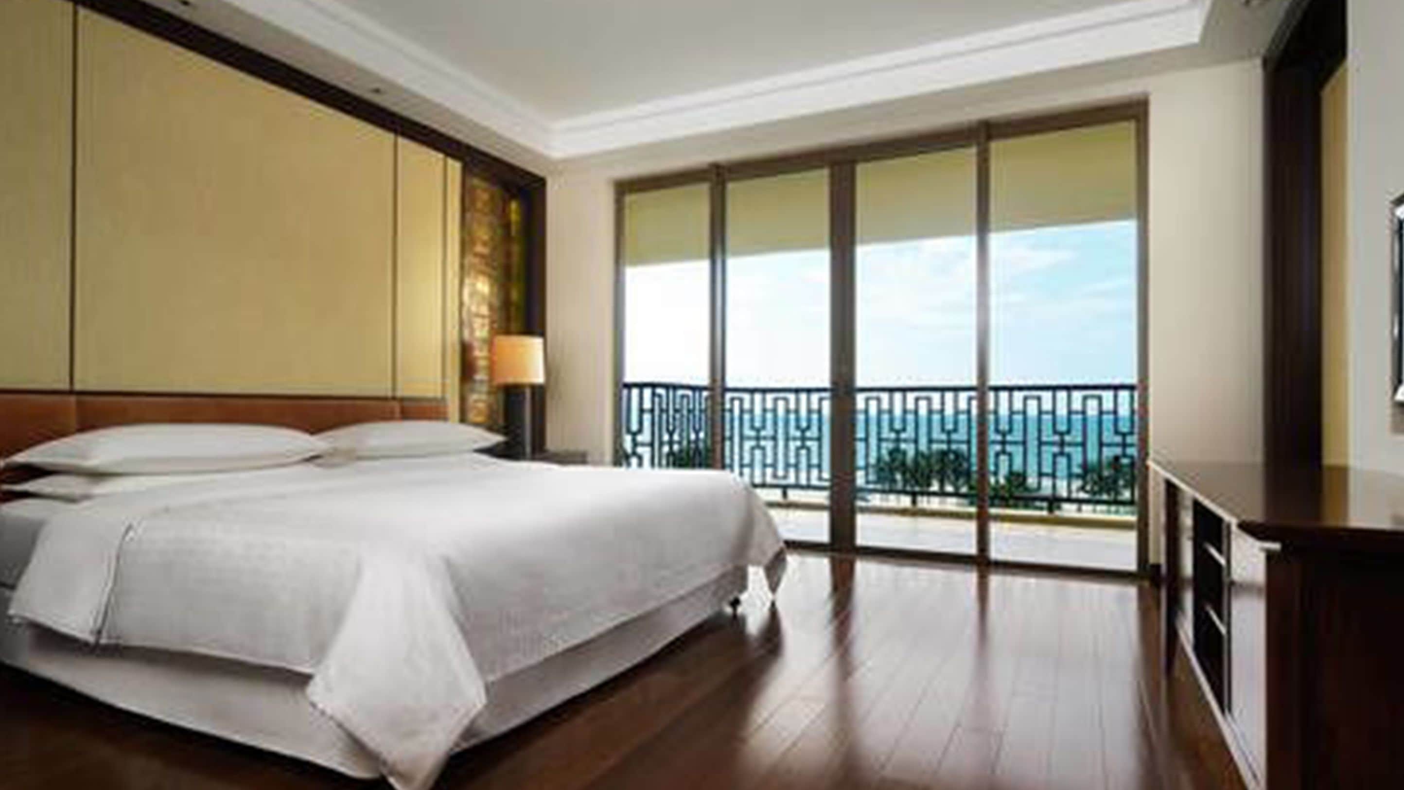 A hotel room with a large bed, a nightstand, a lamp, and a view of the ocean through floor-to-ceiling windows.