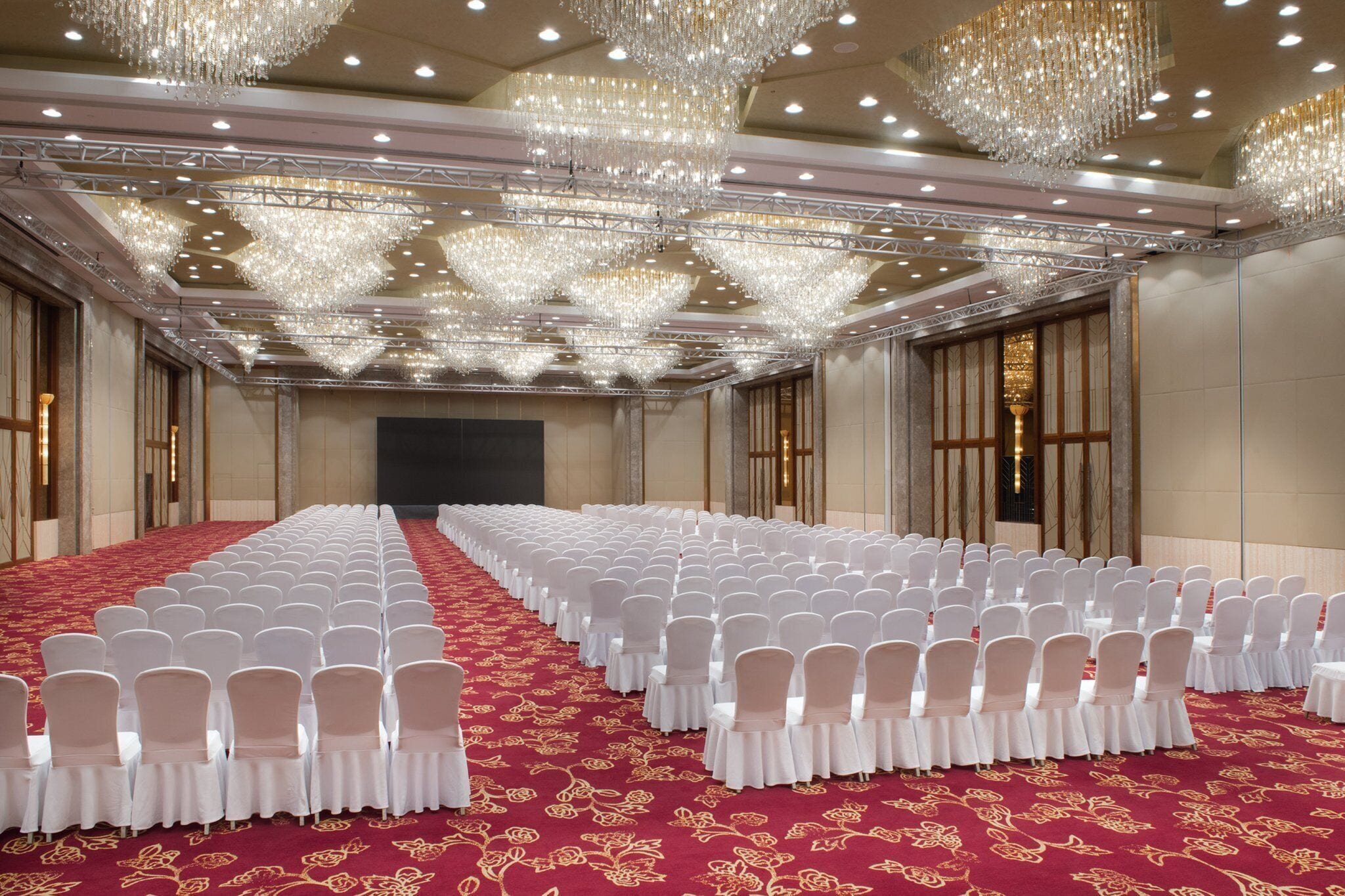 Uma grande sala de conferências com fileiras de cadeiras brancas, um tapete vermelho e várias luminárias de cristal.