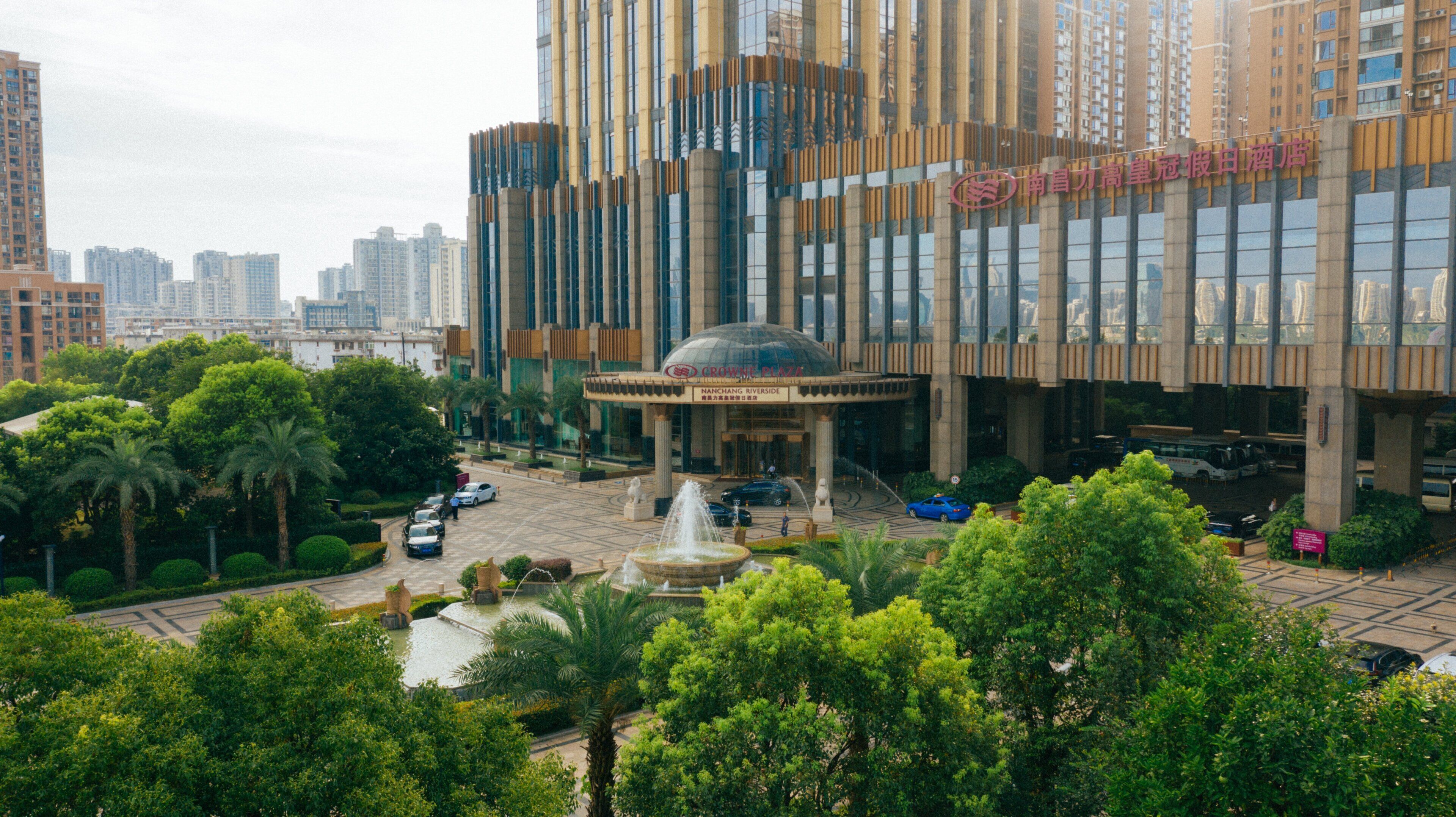 Un hotel moderno con una fuente central, rodeado de exuberante vegetación y rascacielos al fondo.