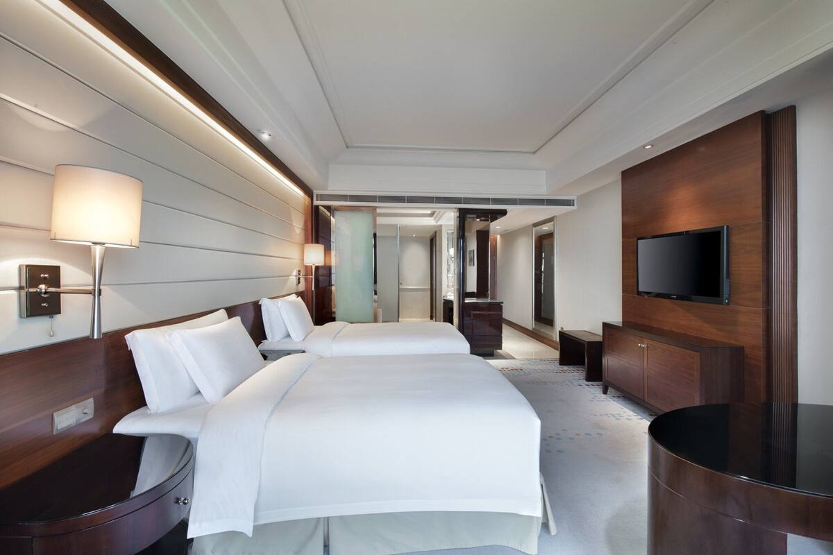 Quarto de hotel com duas camas, uma televisão, um armário de madeira e um espelho grande.
