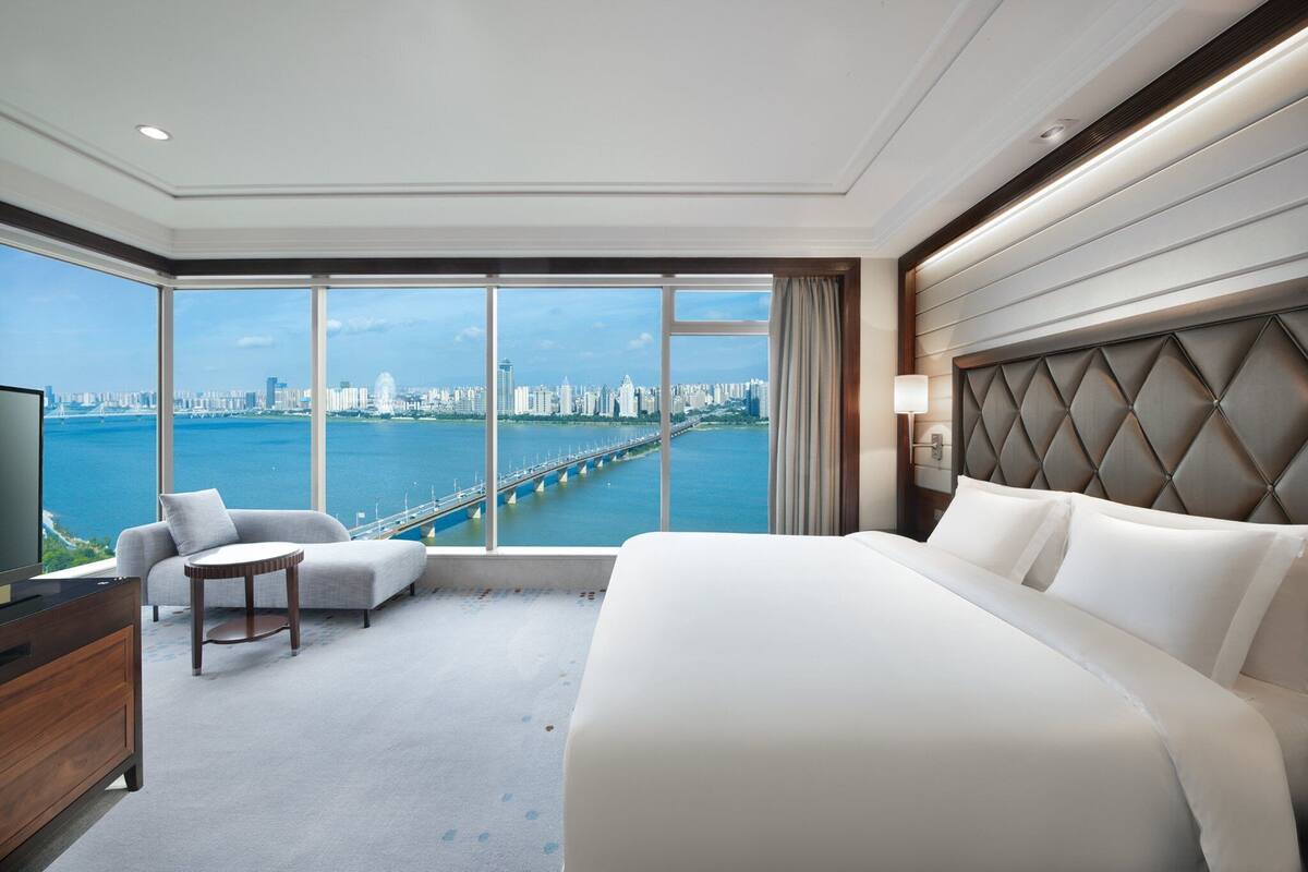 A hotel room with a large bed, a sofa, a TV, and a view of a city skyline and bridge.
