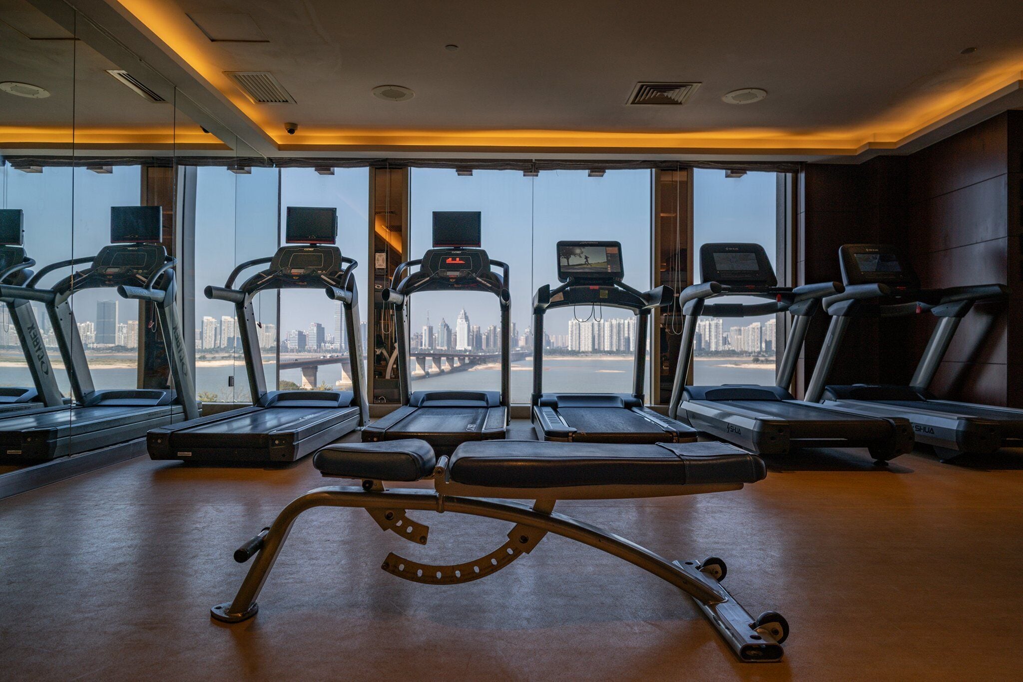 A gym with treadmills and a rowing machine, large windows with a city view, and a wooden floor.