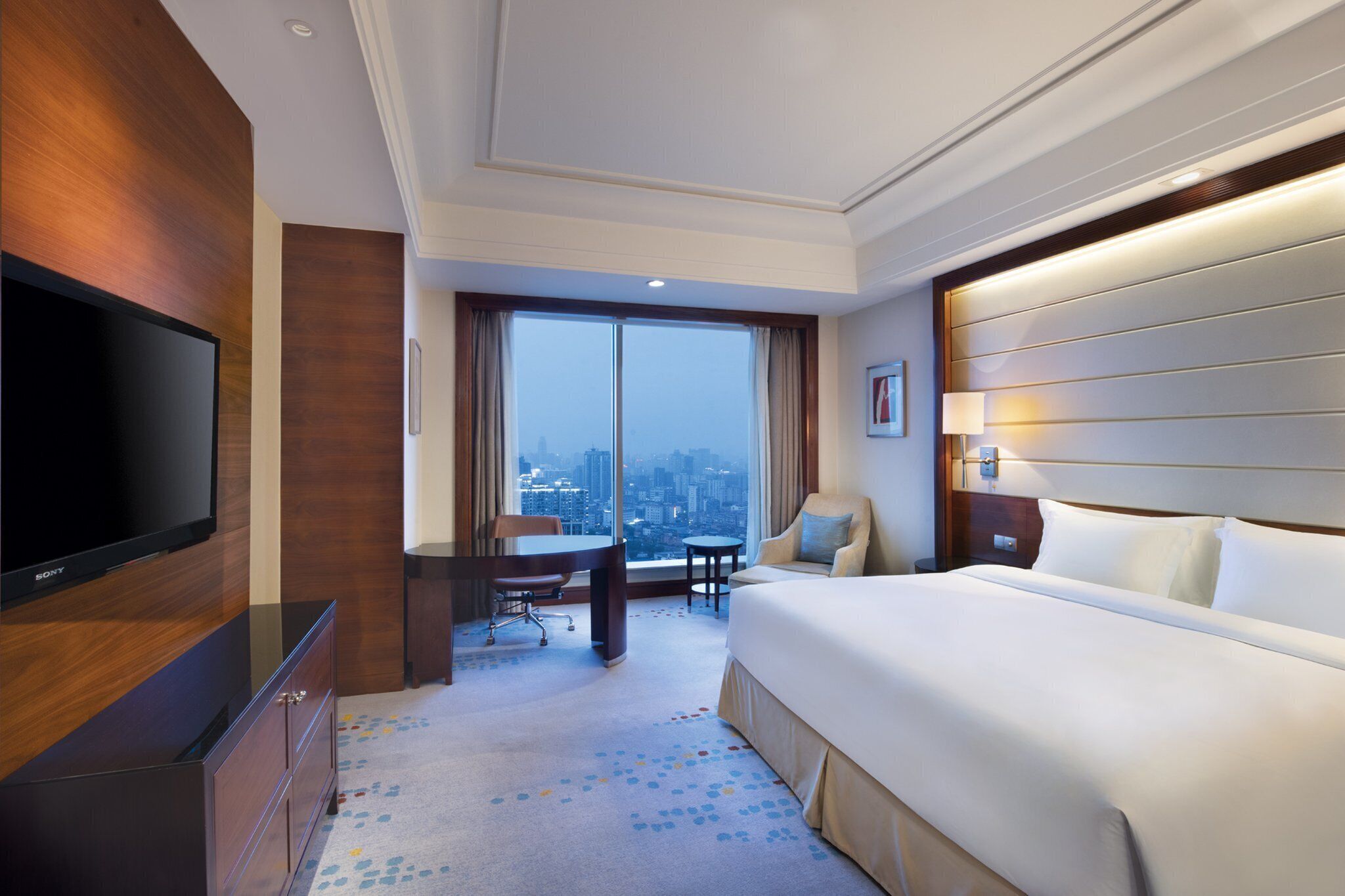 A hotel room with a large bed, a flat-screen TV, a desk, and a chair. There is a large window with a city view.